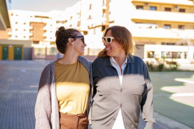 İki neşeli kadın mutluluğu ve bağlantıyı ifade ediyor, günlük kıyafetler giyiyorlar, güneşli bir günde açık havada yürüyorlar.