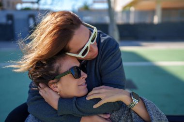 Açık hava sahasında birbirine gülümseyen ve sarılan iki kadın, sakin bir günde iletişim ve zevk gösteriyorlar.