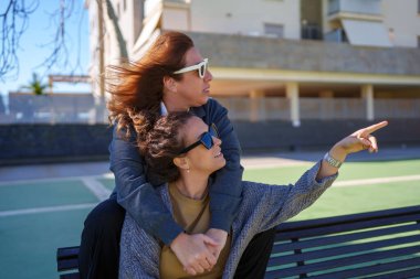 Güneş gözlüğü takan iki kadın bankta sarılıyor, biri işaret ediyor, ikisi de başka tarafa bakıyor. Birlikteliği ve keşfi kucaklamak