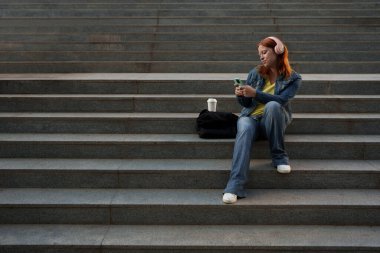 Şehir basamaklarında oturan, pembe kulaklıklı müzik dinleyen ve akıllı telefonuyla etkileşime giren genç bir kadın. Modern dijital yaşam tarzı