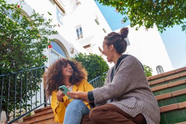 İki mutlu genç bayan arkadaş dışarıda keyifli bir an paylaşıyorlar, akıllı telefondan içeriklerini paylaşıyorlar ve birlikte gülüyorlar.