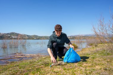 Genç adam aktif olarak göl kıyısındaki plastik şişeleri temizliyor. Çevre gönüllüsü doğa koruma ve atık yönetimi için çalışıyor