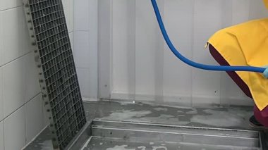 Sarı bir koruyucu giysi ve eldiven giyen biri endüstriyel bir ortamda beton zemini ve metal ızgarayı temizliyor, muhtemelen gıda işleme tesisi. Mavi bir sprey kullanıyor..