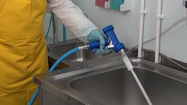 Mavi bir hortuma bağlı yüksek basınçlı bir dezenfektan kullanıyor. İş yerindeki paslanmaz çelik lavaboyu temizlemek ve sterilize etmek için kullanılıyor. Hijyen için kişisel koruyucu giysiler giyiyorlar..
