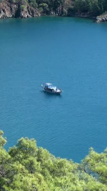 A charming boat drifts peacefully on the vibrant blue Aegean Sea, nestled by verdant hills of Oludeniz, Fethiye, Turkey. Babadag Mountain oversees this serene, sunlit coastal paradise.