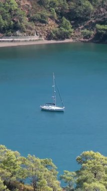Yalnız bir yelkenli, Ege Denizi 'nin ışıldayan mavi sularında zarafetle dinlenir. Fethiye ve Oludeniz yakınlarındaki Babadag Dağı 'nın bir parçası olan yemyeşil tepeler, bu huzurlu gündüz manzarasını çerçeveliyor..