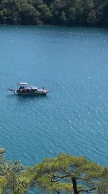 Güzel bir tekne Ege Denizi 'nin canlı turkuaz sularında zarifçe süzülüyor. Babadag Dağı heybetli bir şekilde yüksekte duruyor, gündüz boyunca bu sakin Fethiye, Oludeniz manzarasını çerçeveliyor..