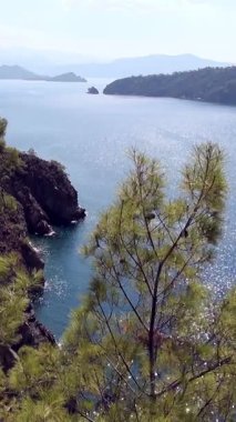 Fethiye Oludeniz yakınlarındaki Ege Denizi 'nin nefes kesici güzelliğine tanık olun. Bereketli yeşil kıyı tepeleri ve canlı çam ağaçları çerçeveli sakin mavi sularda güneş ışığı parlıyor..
