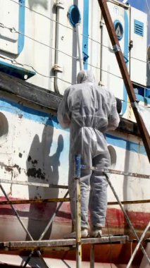 Kendini işine adamış işçiler, geminin gövdesinden eski boya katmanlarını özenle temizler, kolay erişim için iskeleyi kullanır. Parlak güneş ışığı, canlılığını geri kazanma çabalarını vurguluyor..