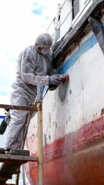 İşçi titizlikle geminin gövdesindeki eski boya katmanlarını özel aletlerle söker ve yeniden boyamak için pürüzsüz bir yüzey sağlar. Açık ve güneşli bir günde marina hareketlilikle dolup taşıyor..