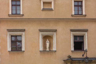 Eski bir hardal evin ön cephesinde dua eden bir rahibenin heykeli, üzerinde beton platformlar ve oymalar olan bordo ahşap çerçeveli pencereler.