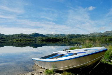 Bir gölün kumlu sahilinde, uzaktaki dağları olan gümüş bir alüminyum tekne. Norveç 'te güzel güneşli bir sonbahar günü ünlü Trolltunga şehir simgesine doğru yol alıyor..