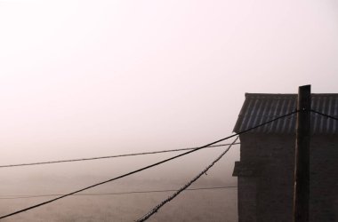 Ön planda elektrik hatları olan sislerle kısmen gizlenmiş kırsal bir binanın bulunduğu sisli bir arazi.
