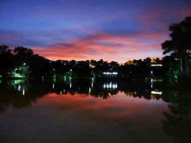 Durgun suda pembe ve mor renkleri olan çarpıcı bir alacakaranlık gökyüzü yansıyor, siluetli ağaçlar..