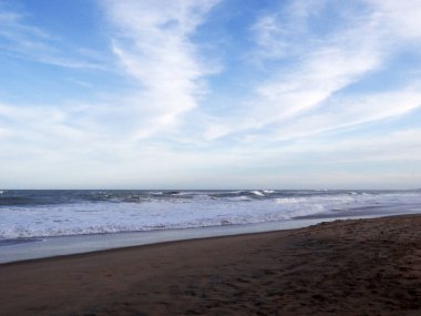 Köpüklü okyanus dalgaları ve ince bulutlarla dolu gökyüzü ile kumlu bir sahilin geniş bir görüntüsü..