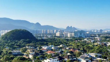 Panoramik bir manzara, yemyeşil tepelerin ve açık mavi gökyüzünün altındaki uzak dağların arasında yuva yapmış canlı bir şehri gösteriyor..