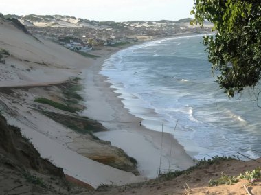 Okyanusa doğru uzanan kum tepeleri olan kumlu bir sahil manzarası. Dalgalar kıyıya vurur parlak bir gökyüzünün altında.