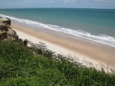 Çimenli bir uçurumdan geniş bir kumlu plaja kadar sakin bir manzara. Hafif dalgalar parlak bir gökyüzünün altında kıyıya vuruyor..