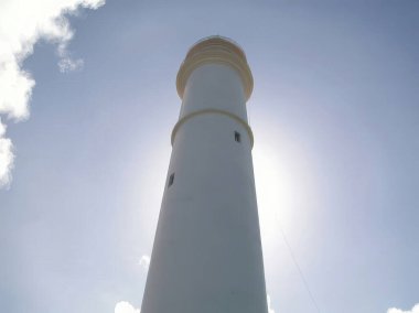 Uzun, beyaz bir deniz feneri, dağınık bulutlu, parlak, güneşli bir gökyüzüne karşı duruyor. Güneş tam olarak binanın arkasında..