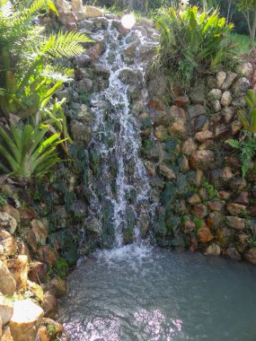 Canlı, çok katmanlı bir şelale, yoğun yeşil yapraklarla çevrili kayalık bir setin üzerinden akar..