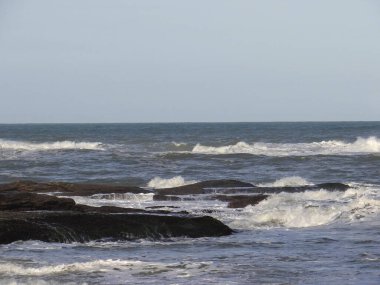 Hafif okyanus dalgaları, solgun bir gökyüzünün altında karanlık, ıslak kayalara karşı yuvarlanıp kırılıyor..