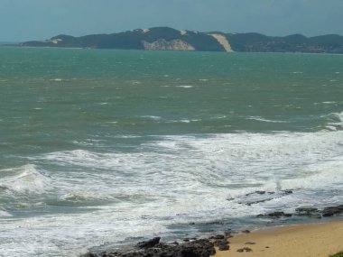 Okyanusun manzarası. Köpüklü dalgalar kumlu bir sahile yuvarlanıyor ve uzaktaki tepeler yuvarlanıyor..