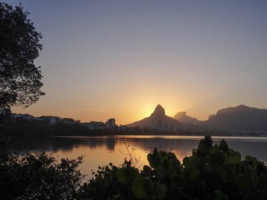A serene sunset casts a warm glow over a calm lake, reflecting mountains and trees.