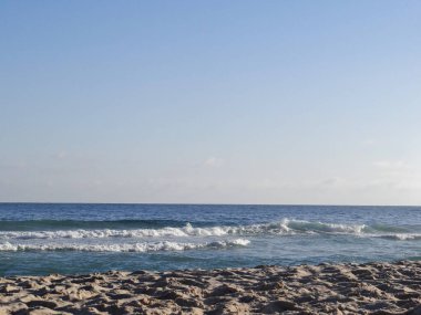 Sakin bir sahil sahnesi, üzerinde hafif dalgalar olan açık mavi bir gökyüzünün altında kıyıya doğru yuvarlanan..