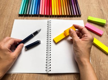Bir insanın ellerinin yukarıdan aşağıya, boş bir spiral deftere sarı yapışkan bir not yerleştirirken görüntüsü. Renkli kalemlerden oluşan tam bir spektrum, sıcak ahşap bir masa arkasında düzgünce dizilmiştir..