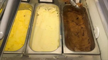 Plastik eldiven giyen biri, yanında vanilya, mango ve muz aromalı metal servis kutusundan zengin çikolatalı dondurmayı tatlı ya da dondurucuya koyar..