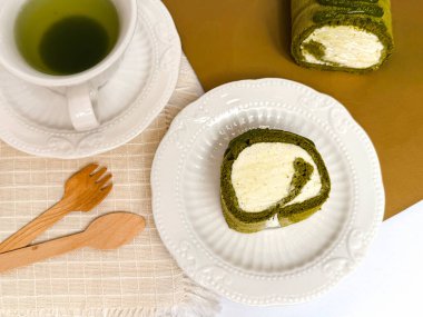 Bir dilim kibritli kek, yeşil çay ve bej bir giysi üzerinde ahşap çatal bıçakla birlikte beyaz bir seramik tabakta servis ediliyor. Tatlı menüleri ve fırın promosyonları için parlak, temiz masa üstü fotoğrafçılığı ideal..