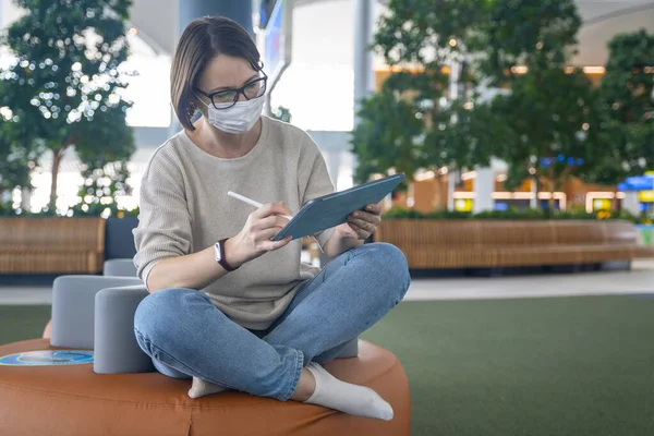 Junge Freiberuflerinnen mit medizinischer Schutzmaske sitzen in Lotus-Position im Business Center oder in der Flughafenlounge und arbeiten mit digitalem Bleistift und Tablet. Leben während einer Pandemie Stockfoto