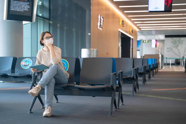 Woman in protective mask and with digital tablet is sitting in airport lounge ready to travel or meet arrived friends. She was distracted from work or social media to listen to departure notification — Stock Photo, Image