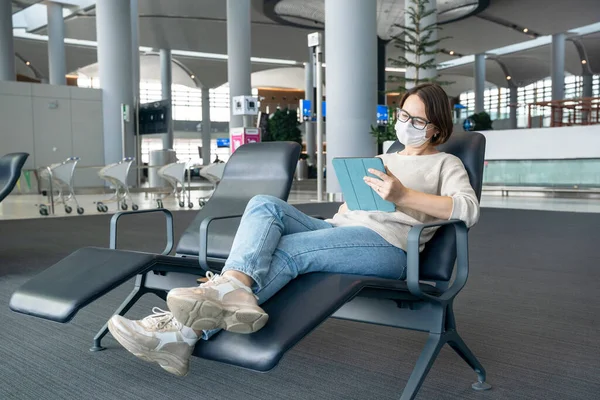 Moderna joven freelancer en máscara de protección médica se sienta cómodamente en sillas y trabaja o crea un nuevo proyecto utilizando lápiz digital y tableta en la sala de espera o en el salón del centro de negocios —  Fotos de Stock