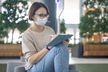 Genç, modern bir kadın halka açık alanda elinde tabletle oturuyor. Günlük kıyafet, yüzünde hijyenik maske. Salgın durumunda herhangi bir yerde çalışma veya yaratıcılık kavramı