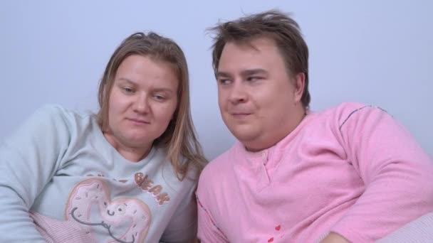 パジャマを着た夫婦はベッドに横になり、子供や他の誰かの話を聞きます。彼らはお互いに疑問を持ち、起こっていることの不条理を笑い始めます。. — ストック動画