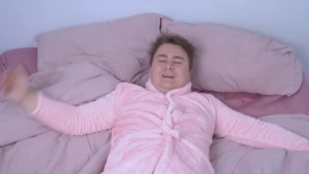 Mladý šťastný muž v legračním růžovém pyžamu s opaskem padl na postel připraven spát a relaxovat po dlouhém těžkém dni objímání svého milovaného jezevčíka psa. Pet doprovází svého majitele všude. — Stock video