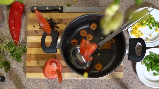 Taze fasulye ve dilimlenmiş sebzeler çorba ya da vejetaryen güveci yapmak için bir kap suya dökülüyor. Yemek tarifi, bıçak, baharat ve kesim tahtası malzemeleri masa yüzeyinde yatıyor.. — Stok video