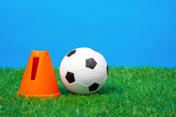 Soporte de retención de cono de plástico una pequeña pelota de fútbol de juguete en la hierba verde del césped artificial, fondo azul, vista frontal, primer plano. — Foto de Stock