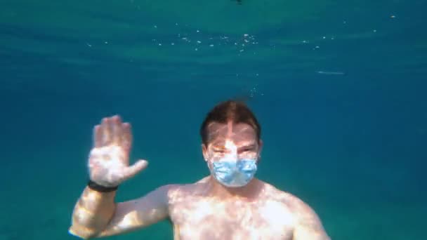 Giovane uomo che indossa maschera medica protettiva sul viso e orologio intelligente al polso è immersioni, nuoto sott'acqua e salutando agitando. Nuove realtà della vita durante la pandemia. — Video Stock