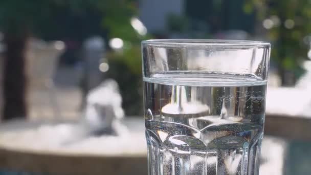 Glass filled with clean drinking water stands on table in cafe or on terrace on warm sunny day, blurred background with decorative fountains create refreshing and cooling oasis in urban landscape. — Stock videók