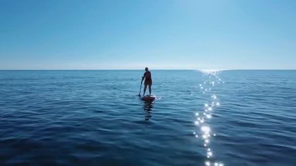 Young woman in cap and sunglasses is rowing oar while standing on SUP board. Active recreation and healthy lifestyle. Shooting with drone moving away from person — Vídeo de Stock