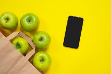 Akıllı telefonun yanındaki parlak sarı yüzey üzerindeki karton paketten çıkan yeşil elmalar. Organik malzeme, ekolojik sağlıklı konsept. İnternetten yiyecek siparişi, eve servis..