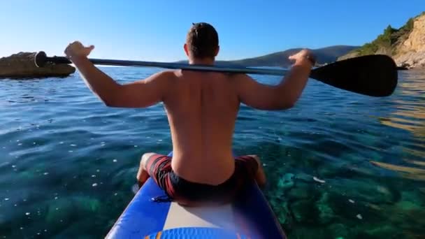 Young man chills and basks in the sun floating on SUP board with paddle thrown around his neck. Guy takes oar in hands and rowing forward to adventure — 비디오