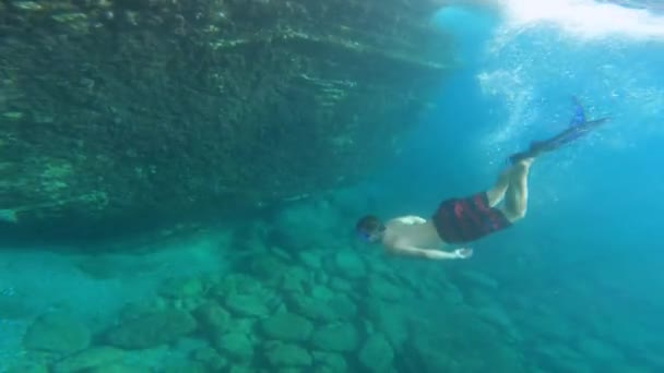 若い男がフリッパーとマスクでシュノーケリングに飛び込みます。男は岩の海の床を探検岩礁に沿って水中の彼の軸上にひっくり返り、アクションカメラで撮影します — ストック動画