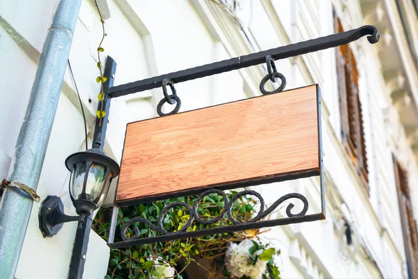 Empty wooden sign with wrought-iron frame and vintage street lamp hangs on the wall of house outside. Blank for advertising or signage of store, cafe, or public organization