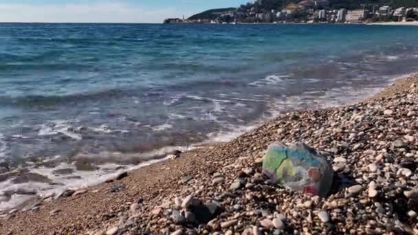 Corazón con fondo de arco iris está pintado sobre piedra que yace en la playa. Las olas de espuma lavan la orilla del mar. Paisaje tranquilo y tranquilo con mar y montaña durante el claro día soleado — Vídeo de stock