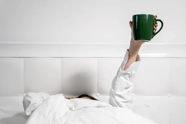Человек в белой рубашке только что проснулся и лежит под теплым одеялом в постели с кружкой кофе в воздухе. Тяжело вставать рано утром. Ежедневно — стоковое фото