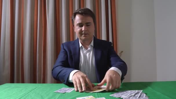 Homem jovem vestindo terno azul oficial sentado da mesa de poker, empurra o dinheiro para a frente, as cartas de jogo estão em todos os lugares. Conceito de paixão do jogador — Vídeo de Stock