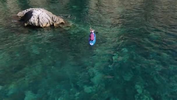 Jong atletisch koppel zit samen in het SUP bestuur. Vrouw peddelt en zwemt rond enorme rots in zee, luie man rust, schieten met drone van hoogte. Gezonde en actieve levensstijl — Stockvideo